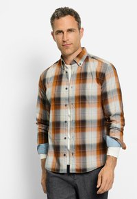 Mann in einem karierten Button-Up-Hemd in Braun, Weiß und Blau über einem weißen Langarmshirt mit dunklen Hosen, stehend vor einem weißen Hintergrund.