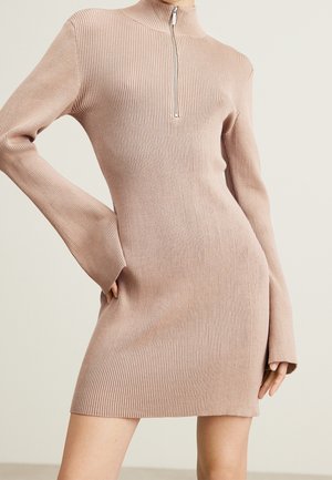 Robe tricotée à côtes en rose clair avec un col montant et une fermeture éclair demi-longueur. Présente des manches longues évasées et une silhouette ajustée avec une fente sur le côté.