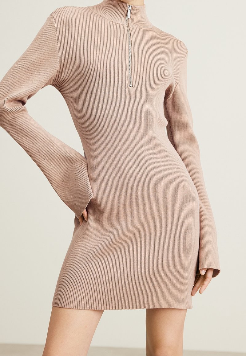Rippstrickkleid in Hellrosa mit hohem Kragen und halblanger Reißverschlussöffnung. Mit langen, ausgestelltäts Ärmeln und einer figurbetonten Silhouette mit einem seitlichen Schlitz.
