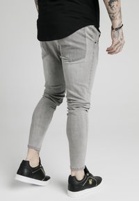 Jeans de mezclilla gris ajustados con bolsillos traseros y rodillas texturizadas, combinados con zapatillas negras y blancas con detalles dorados.