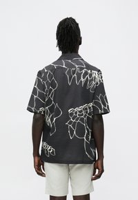 Homem com dreadlocks escuros visto de costas, vestindo uma camisa preta de manga curta com padrões abstratos de linhas brancas e calções bege claro.