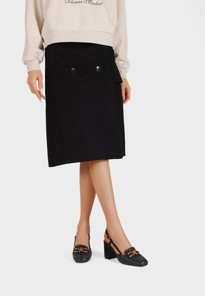 Gonna nera al ginocchio con doppio bottone frontale, abbinata a scarpe slingback nere con texture e dettagli dorati.