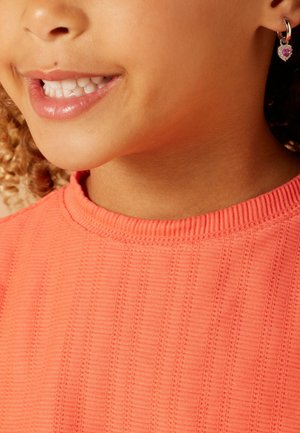 Haut texturé corail avec encolure côtelée ; présente une boucle d'oreille en forme de cœur avec un accent rose. Peau lisse avec un sourire.