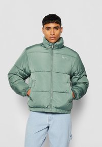 Chaqueta acolchada verde con un alto cuello, con cierre de cremallera, puños elásticos y paneles cosidos. Textura suave con detalle del logo.