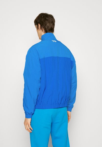 Két tónusú kék, könnyű széldzsekit viselő személy, rugalmas mandzsettákkal és alsó peremmel, egy sima fehér háttér előtt áll.