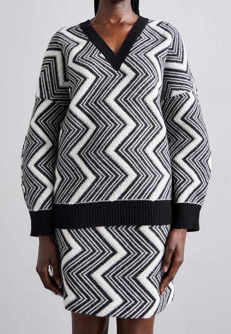 Pull à manches longues col en V à motif zigzag noir et blanc et jupe assortie sur une personne sur un fond uni.