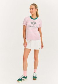 Kobieta stoi ubrana w różową koszulę w paski z napisem "Checked In", białą spódnicę, różowe skarpetki i zielone trampki na jednolitym białym tle.