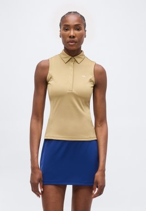 Femme portant un haut beige sans manches à col et une jupe bleue, debout avec une expression neutre sur un fond uni.