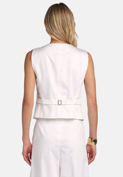 Blondínka v nosení bieleho bezrukávového vestu s nastaviteľnou prackou vzadu a bielymi nohavicami na mieru, zlatý náramok na pravom zápästí.