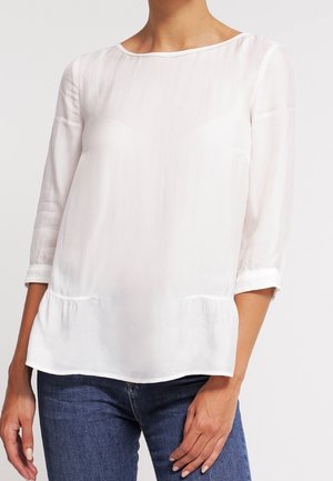 Blouse blanche semi-transparente avec un décolleté arrondi et des manches trois-quarts, présentant un ourlet peplum subtil et une texture lisse.