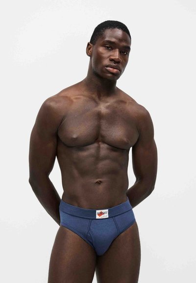 Homme musclé debout devant un fond blanc, portant un slip bleu avec un patch logo sur la ceinture, les mains derrière le dos.