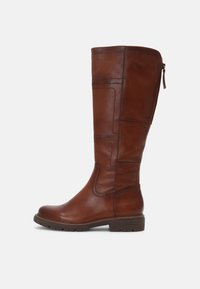 Botte en cuir marron jusqu'au genou avec des détails de panneaux cousus, un petit talon empilé et une fermeture éclair à l'arrière sur fond blanc.