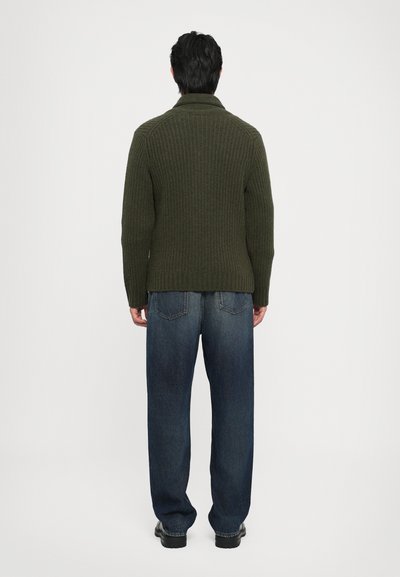 Pulover ribbed de culoare verde închis, cu guler înalt, asortat cu blugi lejeri din denim albastru. Vederea din spate subliniază un design simplu.