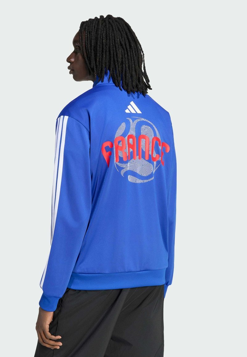 Personne aux cheveux tressés portant une veste Adidas bleue avec des bandes blanches et le texte rouge « FRANCE » au-dessus d'un graphique de ballon à motifs dans le dos.
