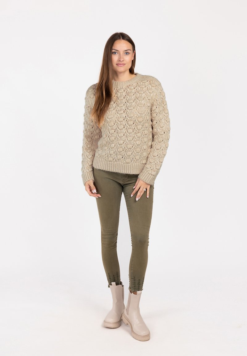 Beige gestrickter Pullover mit einem strukturierten Muster, grüne eng anliegende Hose und hellbeige Ankle Boots. Das Outfit hat gerippte Bündchen und Saum.