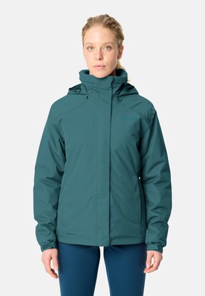 Vrouw draagt een turquoise waterdichte jas met capuchon en marineblauwe legging, staand voor een effen lichte achtergrond.
