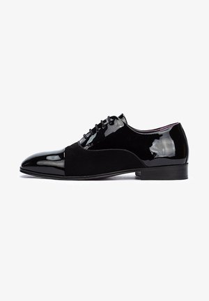 Zapato de vestir negro que combina charol brillante y gamuza mate, con punta afilada y cierre de cordones. Tacón plano.