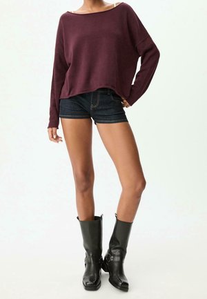 Femme portant un pull bordeaux ample, un short en jean foncé et des bottes en cuir noires mi-mollet, debout devant un fond blanc uni.