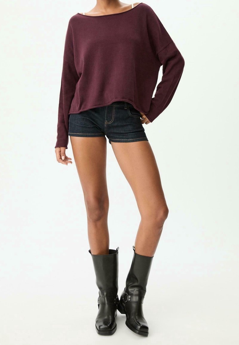 Femme portant un pull bordeaux ample, un short en jean foncé et des bottes en cuir noires mi-mollet, debout devant un fond blanc uni.