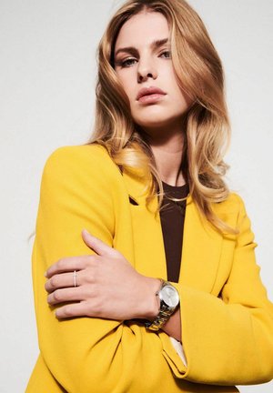 Sølv armbåndsur med rund skive og led-bånd, båret på en hånd med en ring. Modellen har en lys gul skræddersyet blazer på.