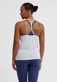 Vitt atletiskt linne med racerback-design, med mörkblå detaljer och en texturerad tyg. Modellen har synliga tatueringar på sina armar.