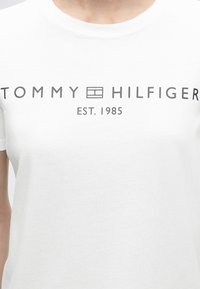 Λευκό μπλουζάκι με μαύρο κείμενο "TOMMY HILFIGER" και λογότυπο στο κέντρο, και το "EST. 1985" τυπωμένο κάτω στο στήθος.