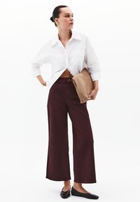 Camicia bianca oversize con dettagli nei bottoni, abbinata a pantaloni larghi color bordeaux. Il modello tiene una pochette marrone e indossa ballerine nere.