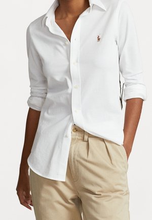 Persona con una camisa blanca abotonada de mangas remangadas y pantalones beige plisados, de pie con una mano en el bolsillo.