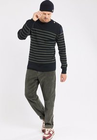 Homme portant un pull rayé noir et vert, un pantalon en velours côtelé gris, un bonnet marine et des baskets rouge et blanc, se tenant contre un fond blanc.