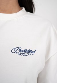 Sweat-shirt blanc à col rond avec texte brodé en bleu marine "Prohibited Racing Team" sur la poitrine, porté par une personne aux cheveux foncés.