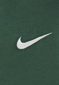 Zaļš kokvilnas audums ar gludu tekstūru, centrā izvietots balts izšūts Nike swoosh logotips.