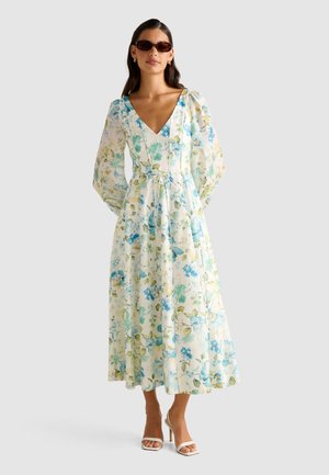 Femme portant une robe midi blanche à fleurs avec des manches longues bouffantes, un décolleté en V, une ceinture à la taille, des lunettes de soleil marron et des sandales à talons blanches.