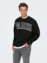 Black knitted sweater with "PARIS" in white letters across the chest, worn over a white shirt, paired with gray jeans.