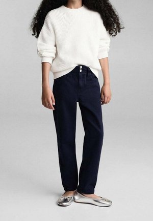 Bambino con capelli ricci che indossa un maglione bianco, pantaloni blu navy a vita alta e ballerine argento, in piedi davanti a uno sfondo semplice.