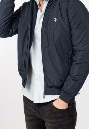 Mężczyzna w granatowej bomberce na jasnoniebieskiej koszuli zapinanej na guziki, z jedną ręką w kieszeni, a drugą poprawiający kołnierz kurtki.