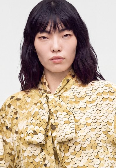 Γυναίκα με μαύρα μαλλιά μέχρι τους ώμους, φοράει χρυσή και μπεζ μπλούζα με κυματιστό μοτίβο και μεγάλο γιακά με φιόγκο.