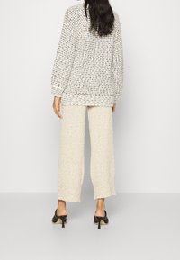 Dicker, cremfarbener Strickpullover mit strukturiertem Muster, kombiniert mit weit geschnittenen, cremfarbenen Hosen. Das Model trägt schwarze, hohe Mules.