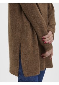 Brązowy, przytulny sweter o miękkiej fakturze, z bocznymi rozcięciami i długimi rękawami. Połączony z ciemnoniebieskimi dżinsami, dłonie delikatnie złączone.