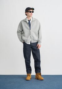 Felpa grigia chiara con cerniera sopra una maglia bianca e cravatta blu, jeans di denim blu e stivali color cuoio con lacci, con un berretto a righe e occhiali da sole.