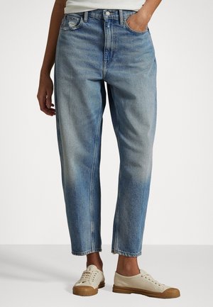 Personne portant un jean bleu clair coupe décontractée et des baskets en toile beige sur un fond blanc uni.