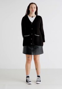 Czarny kardigan z białymi detalami, dekolt w szpic, zapięcie na guziki, zestawiony z czarną skórzaną spódnicą i czarnymi sneakersami na wysokiej podeszwie.