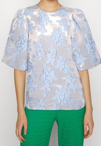 Blusa rosa claro con patrón floral azul texturizado, mangas cortas de globo y cuello redondo. Combinada con pantalones verdes texturizados.