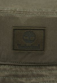 Brunt tekstilpatch med "Timberland" logo og træemblemet, omgivet af grøn syning på en khakibat. Flad, rektangulær form.