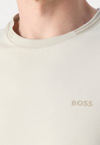 T-shirt beige chiaro con colletto a girocollo a coste, tessuto testurizzato e un discreto logo "BOSS" in rilievo sul lato sinistro del petto.