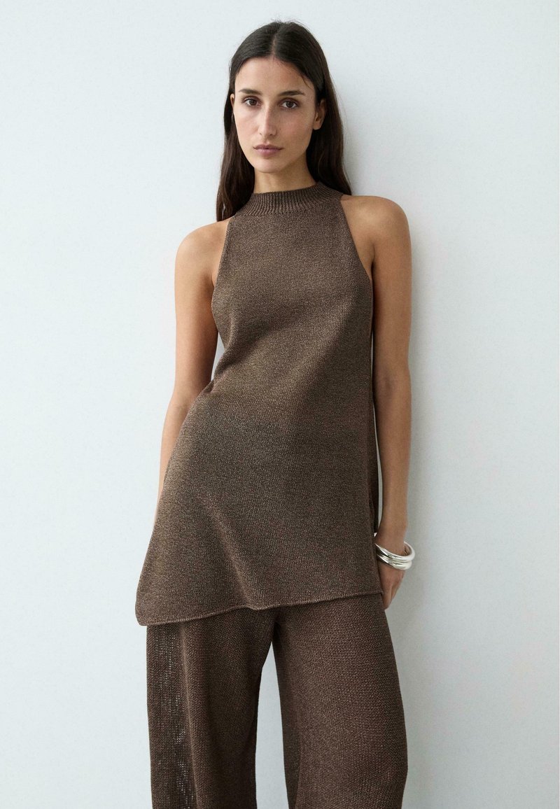 Vrouw met lang donker haar, gekleed in een mouwloos bruin gebreid tuniek en bijpassende wijde broek, staand tegen een eenvoudige lichte achtergrond.
