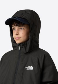 Černá nepromokavá bunda se zapínáním na zip vpředu, nastavitelnou kapucí a bílým logem. Pod kapucí je nošena tmavě modrá baseballová čepice.