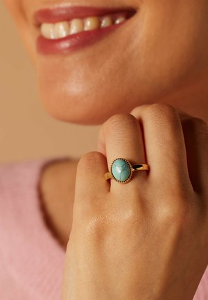 Bague en or avec une pierre ovale en turquoise, entourée d'un bord perlé. Main reposant sur le menton, peau visible dans un éclairage doux.