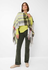 Poncho gris à carreaux avec des accents verts, jaunes et rouges, drapé sur un haut vert vif, associé à un pantalon en velours côtelé vert foncé et des chaussons beiges.