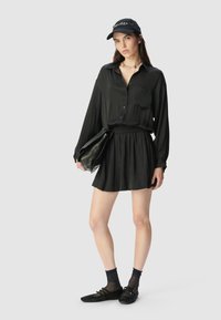 Schwarzhemd mit langen Ärmeln, kombiniert mit einem plissierten schwarzen Rock und einer schwarzen Clutch. Eine schwarze Mütze, Knöchelsocken und flache Schuhe runden das Outfit ab.