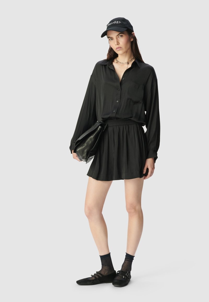 Schwarzhemd mit langen Ärmeln, kombiniert mit einem plissierten schwarzen Rock und einer schwarzen Clutch. Eine schwarze Mütze, Knöchelsocken und flache Schuhe runden das Outfit ab.